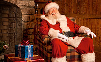 Santa sitting on a chair near a fireplace with a gift
